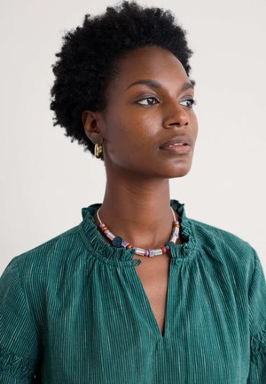 Chemisier teal à rayures avec des manches bouffantes et un col à volants. Agrémenté d'un collier de perles multicolores et de boucles d'oreilles en or.