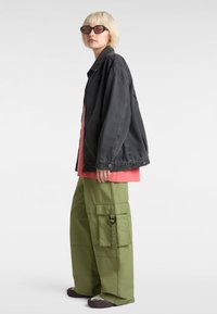 Schwarze Jeansjacke, leuchtendes pinkes Hemd, übergroße grüne Cargo-Hosen mit Taschen und braune gemusterte Schuhe. Schwarze Sonnenbrillen runden den Look ab.