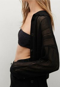 Schwarze, strukturierte, offen gestrickte Strickjacke mit langen Ärmeln und Knöpfen, die über einem dunkel gemusterten Oberteil getragen wird, mit einem cropped Schnitt und lässiger Passform.