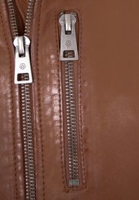 Braune Lederjacke mit zwei Metallreißverschlüssen mit rechteckigen Zügen. Glatte Textur, dezenter Glanz und feine Nähte.