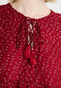 Rochie roșie cu flori, cu plisuri adunate, decolteu în V și închidere cu fundă decorată cu ciucuri. Material moale cu un finisaj texturat și modele delicate.