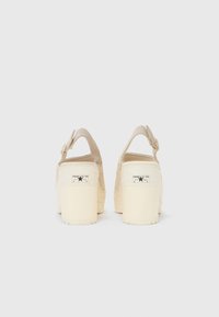 Sandales à plateforme beiges avec un dos ouvert, une sangle ajustable et une semelle en caoutchouc. Présente une étiquette de marque au talon et une texture lisse.