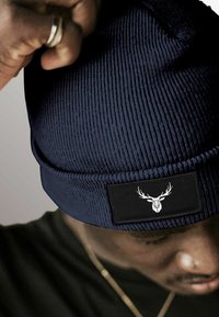 Navy knit beanie with ribbed texture. Features a black patch with a white deer logo. Close-up, partially obscured by a hand.