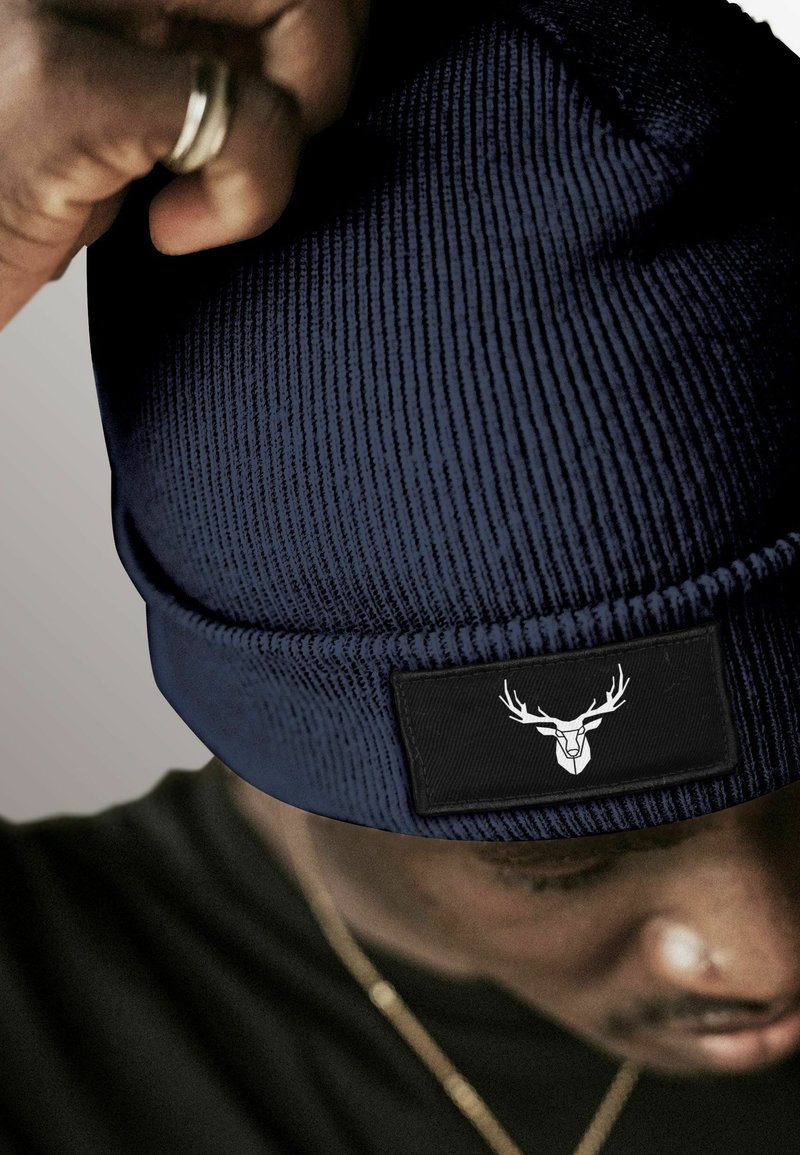 Navy knit beanie with ribbed texture. Features a black patch with a white deer logo. Close-up, partially obscured by a hand.