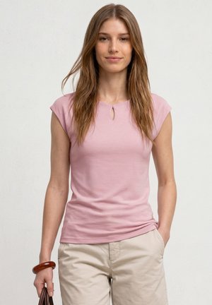 Jeune femme aux longs cheveux bruns porte un haut rose à manches courtes et un pantalon beige, tenant un sac à main marron avec une main dans la poche.