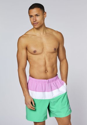 Oberkörperfreier Mann steht mit einer Hand in der Tasche einer pastellrosa, weißen und grünen Badehose, neutraler Hintergrund.
