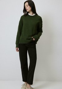 Femme debout portant un pull vert foncé sur une chemise blanche à col, un pantalon noir et des baskets beige, sur un fond clair uni.