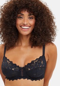 Mujer sonriente con cabello rizado, vistiendo un sujetador negro de encaje con patrones florales y tirantes delgados, sobre un fondo liso.