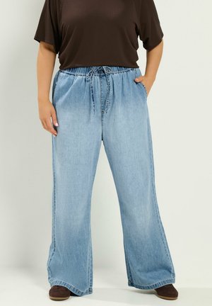 Person trägt hellblaue Jeans mit weitem Bein und Kordelzug in der Taille sowie dunkelbraune Schuhe, steht mit einer Hand in der Tasche.
