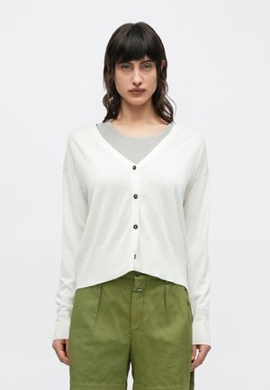 Kvinde med mørkt hår iført en hvid knappet cardigan over en grå top og olivengrønne bukser, stående mod en ensfarvet baggrund.