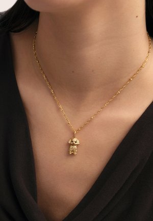 Collar de cadena de oro con un pequeño colgante de oro en forma de perro texturizado, llevado alrededor del cuello de una persona sobre una camiseta de cuello en V oscura.