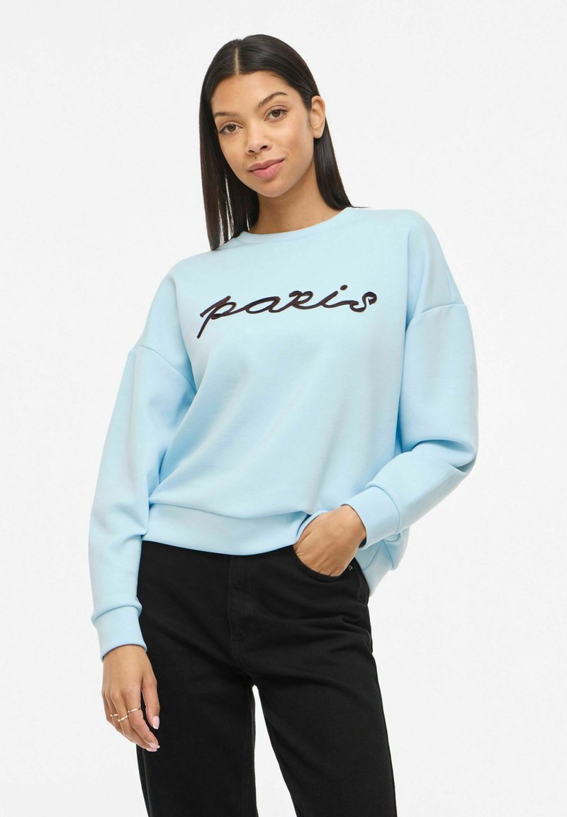 Jeune femme aux cheveux noirs et raides portant un sweat-shirt bleu clair avec le texte "paris" et un pantalon noir, debout avec une main dans la poche.