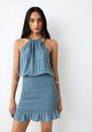 Femme aux longs cheveux noirs portant un haut dos nu bleu à motif et une mini-jupe à volants assortie, debout devant un fond blanc.