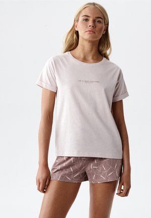 Blondynka w jasnoróżowej koszulce z podwiniętymi rękawami z napisem oraz w mauve krótkich spodenkach z białym abstrakcyjnym wzorem, stoi na białym tle.