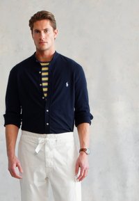 Camisa azul marinho de botão com colarinho, usada sobre uma t-shirt listrada em amarelo e azul marinho, combinada com calças brancas de cordão.
