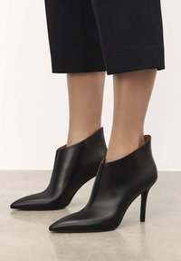 Bottines en cuir noir avec un bout pointu, un talon aiguille haut et un design découpé élégant au-dessus de la cheville, avec une texture lisse.