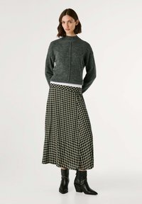 Maglione grigio a maglia con collo alto, abbinato a una gonna lunga a quadri con un motivo a griglia bianco e nero. Stivali neri alla caviglia completano l'outfit.