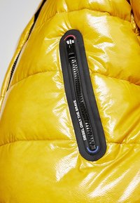 Giacca a imbottitura gialla con finitura lucida, con una tasca con cerniera nera sul braccio e testo del marchio lungo la cerniera.