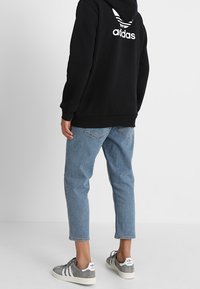 Person klädd i svart Adidas-hoodie, avklippta ljusblå jeans och grå Adidas-sneakers, som står mot en enfärgad ljus bakgrund.