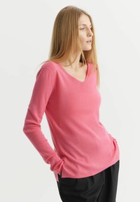 Sweter z dekoltem w serek i długim rękawem w jasnoróżowym kolorze, wykonany z miękkiego materiału, o luźnym kroju i z bocznymi kieszeniami, w zestawie z czarnymi spodniami.