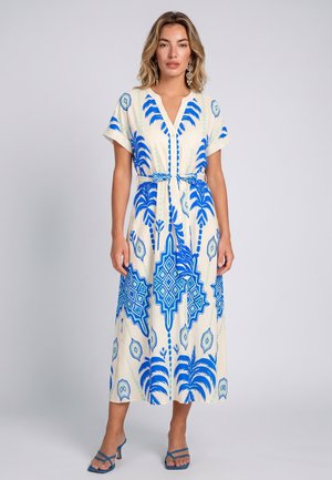 Frau trägt ein weißes Midikleid mit blauen geometrischen und Palmenblattmustern, einem Taillengürtel, kurzen Ärmeln und blauen Sandalen mit Absatz.