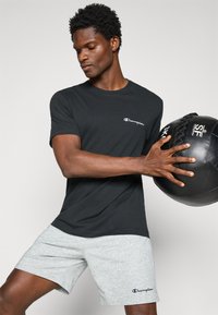 Schwarzes T-Shirt mit kurzen Ärmeln und dem weißen "Champion"-Logo. Graue Sportshorts mit Taschen. Das Model hält einen schwarzen Medizinball.