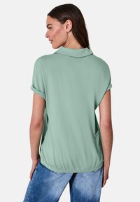 Mintgrünes kurzärmliges Oberteil mit Polo-Kragen, umgeschlagenen Ärmeln und gerafftem Saum. Glatte Textur; lockere Passform. Trägt man mit hellblauen Jeans.