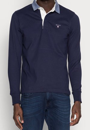 Mand iført en mørkeblå langærmet polo med lyseblå krave og mørkeblå jeans, stående mod en ensfarvet baggrund.