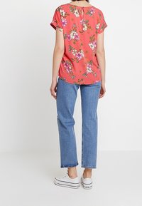 Bluse mit Blumenmuster in Korallenfarbe, kurzen Ärmeln und Knopfverschluss hinten, kombiniert mit hellblauen geraden Jeans und weißen Turnschuhen.
