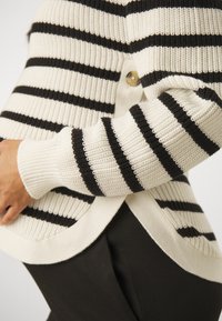 Stripet cardigan med svart og kremfarge, ribbestrikket tekstur, V-hals og knappelukking på siden. Mykt stoff med avslappet passform.