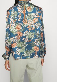 Blouse fleurie bleue avec des motifs orange, blanc et vert ; dotée d'un col à volants et d'une découpe en forme de goutte dans le dos, manches longues avec poignets élastiques.