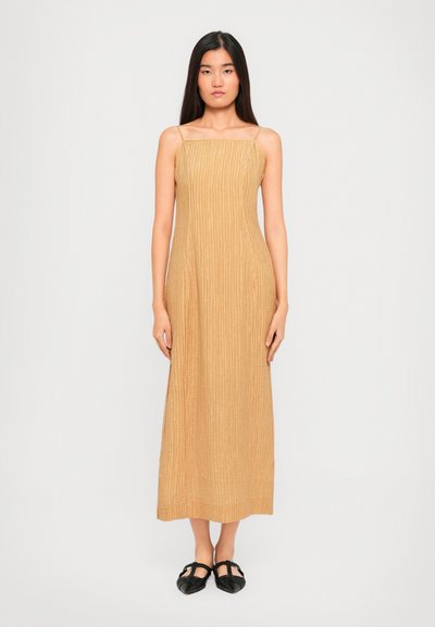 Woman standing wearing a beige textured sleeveless midi dress with thin straps and black pointed-toe flats.