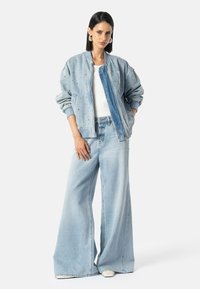 Nő világoskék szegecses farmerdzsekiben, fehér felsőben, világoskék bő szabású farmernadrágban és fehér cipőben, áll a zsebébe tett kezével.