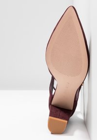 Chaussure à talon haut en daim couleur bordeaux avec un bout pointu et une semelle en caoutchouc texturée. Côtés ouverts et doublure intérieure lisse.