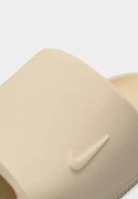 Μπεζ σαγιονάρα με ανάγλυφο λογότυπο Nike στο φαρδύ λουρί, υφή επιφάνειας και εμφανή ραβδώσεις κατά μήκος της άκρης της σόλας.