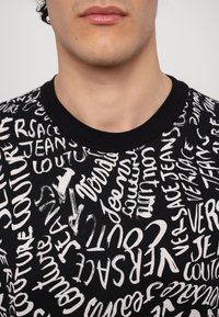 Juodas marškinėlis su baltomis abstrakčiomis tekstinėmis raštais. Su apvalia iškirpte, trumpomis rankovėmis, pagamintas iš minkštos medžiagos. Tekstas apima "Versace Jeans Couture."