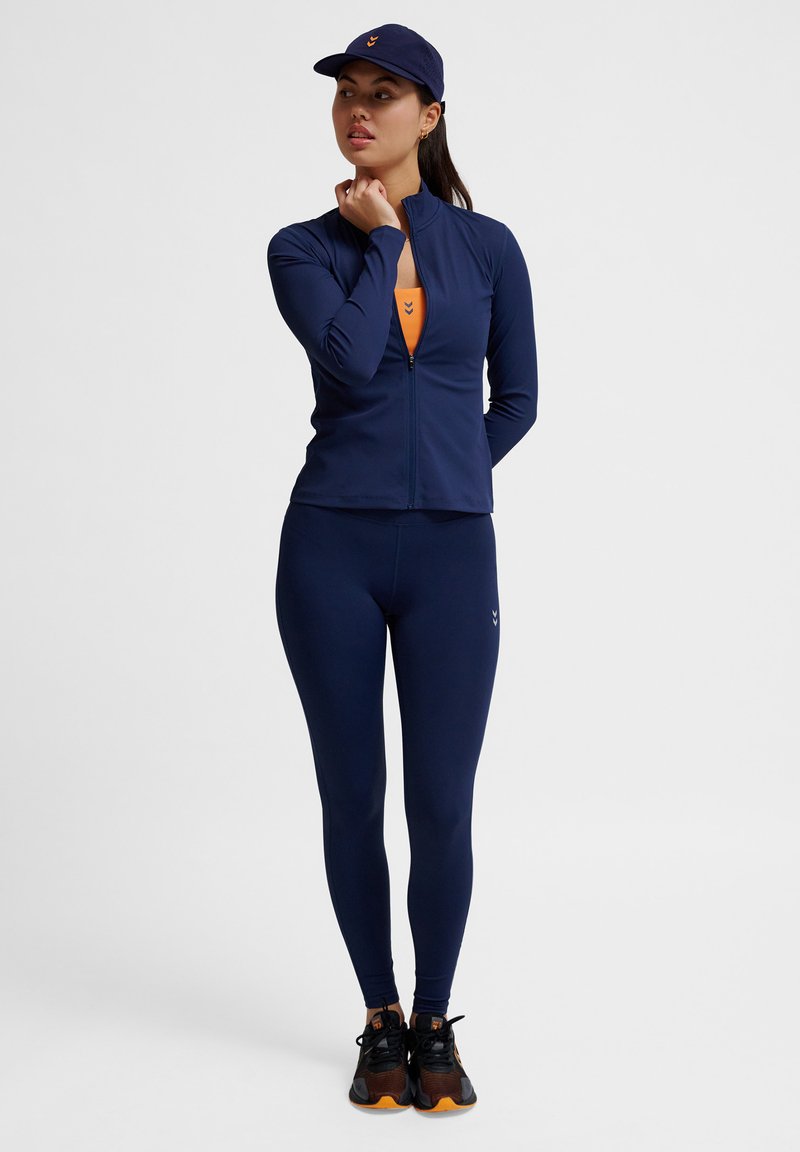 Marinblå idrottsjacka med dragkedja över en orange topp, kombinerad med marinblå leggings. Har en keps med en orange logotyp och svarta sportskor.