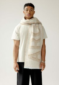 Crème en beigekleurige sjaal van zachte wol met groot "ARC" tekstpatroon, met franjes aan de randen, gedragen om de nek over een licht T-shirt.