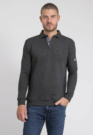 Homme aux cheveux courts et barbe portant un polo à manches longues gris foncé et un jean bleu, debout devant un fond blanc uni.