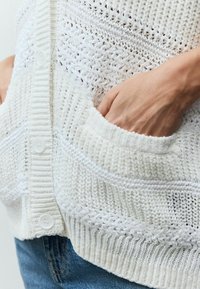 Cardigan blanc en tricot présentant des motifs texturés, des poignets côtelés et des poches frontales ; fermé par des boutons blancs au centre.