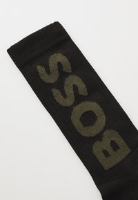 Chaussette noire avec des lettres tissées en olive sur le côté. Le matériau semble doux avec une texture lisse. Le design présente une forme simple et allongée.