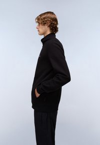 Schwarze Fleece-Zipperjacke mit hohem Kragen, Seitentaschen und gerippten Bündchen. Das Model trägt schwarze Hosen und wird im Profil vor einem hellen Hintergrund gezeigt.