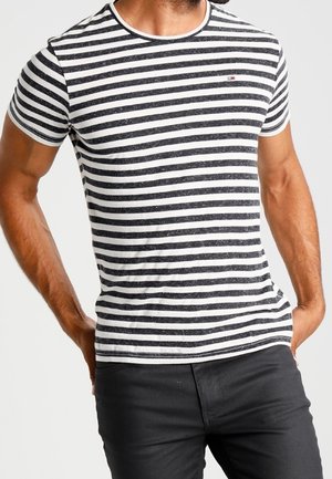 Mand iført en tætsiddende sort-hvid horisontalt striped kortærmet t-shirt og sorte bukser med hænderne hvilende på hofterne.