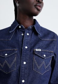 Cămașă din denim cu o spălare de culoare albastră închisă, având nasturi albi cu prindere tip snap, două buzunare pe piept și cusături galbene contrastante într-un model zigzag.