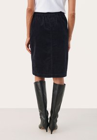 Egy nő térd fölé érő fekete kordbársony szoknyát és fekete sarokkal ellátott, térd magas bőrcsizmát visel, egy sima világos háttér előtt áll.