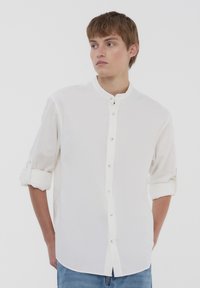 Camicia bianca con colletto alla mandarina, maniche lunghe arrotolate fino ai gomiti e una sottile trama, dotata di bottoni abbinati e orlo dritto.