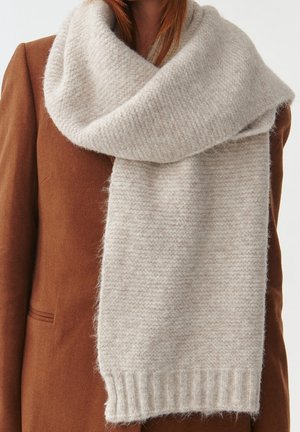 Femme portant une grosse écharpe tricotée gris clair sur un manteau marron, avec l'écharpe lâchement enroulée autour du cou et pendante.