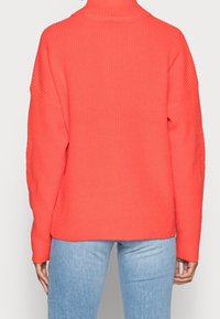 Stickad tröja i klar korallfärg, hög krage, lös passform och ribbade detaljer. Långa ärmar som smalnar av vid handlederna, kombinerat med ljusblå jeans.