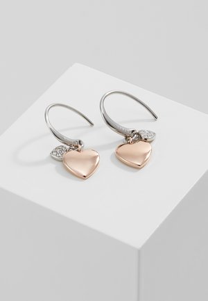 Boucles d'oreilles en ton argent ornées de charms en forme de cœur en or rose et de petits accents de pierres précieuses claires, suspendues à des crochets incurvés sur un présentoir blanc.