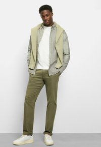 Mann trägt ein hellgrünes Hemd über einem weißen T-Shirt, olivgrüne Hose und weiße Sneakers mit cremefarbenen Akzenten.